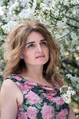 Çiçek desenli uzun saçlı çekici kadının seçici odağı bahçedeki çiçek açan kiraz ağacına bakıyor.