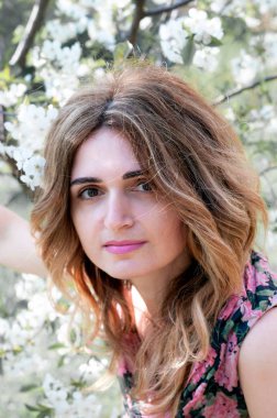 Güzel, kendine güvenen, uzun saçlı bir kadın çiçek açan kiraz ağacının önündeki kameraya bakıyor. Bahçesinde beyaz çiçekler ve güneş ışığı var.