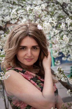 Ağacın yanında, beyaz kiraz dalları açmış, kameraya bakan ve güneşli bahçede uzun saçlarını tutan çekici, gülümseyen bir kadın.