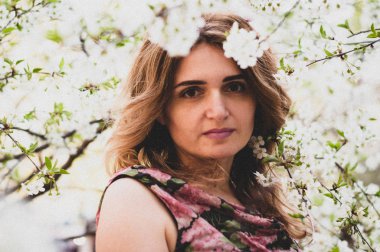 Çekici, uzun saçlı, pembe siyah elbiseli, çiçek desenli beyaz kiraz dallarıyla ağacın yanında duran ve güneşli bahçede kameraya bakan çekici bir kadın.