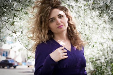 Kendine güveni tam çekici bir kadın mor kazaklı güzel uzun saçlı bahçedeki beyaz çiçeklerle çiçek açan kiraz dallarının önündeki kameraya bakıyor.