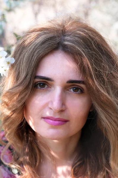 Çekici, kendine güvenen, uzun saçlı, çiçekli bir kadının portresi. Bahçede gün ışığıyla açan dalların önündeki kameraya bakıyor.