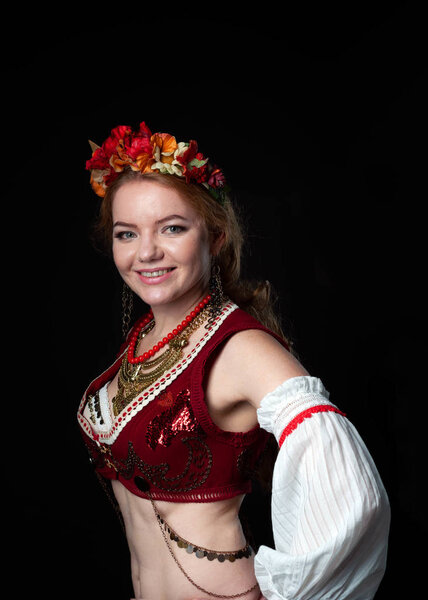 portrait of beautiful smiling woman in traditional costume in ukranian national wreath with flowers, red vest and golden necklace looking at camera isolated on black