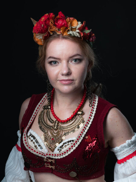 portrait of attractive confident woman in traditional costume in ukranian national stile with circlet of flowers, red vest and golden necklace looking at camera isolated on black
