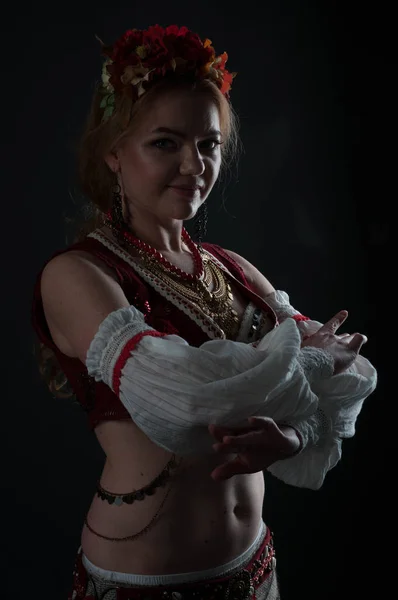 low-key portrait with back light of beautiful woman in traditional folk slavic costume for belly dance holding hands near chest and looking at camera isolated on black