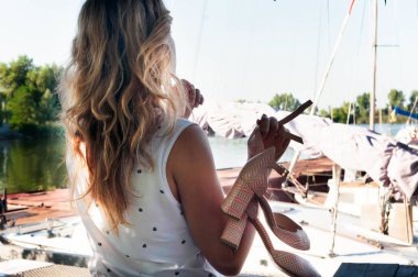 Beyaz bluz giyen, elinde ayakkabılar olan ve nehir kıyısındaki teknelerin önünde gün ışığıyla aydınlanan bir kadının arka görüntüsü.