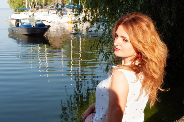 side view of beautiful woman with long hair in white blouse sitting near river with boats lighted with sunshine and looking at camera in yachts club