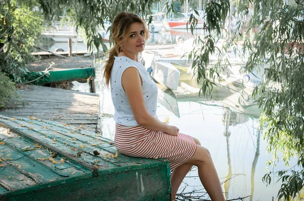 side view of woman in white blouse with ponytail sitting on old wooden green boat and looking at camera in front of tree branches near river with yachts