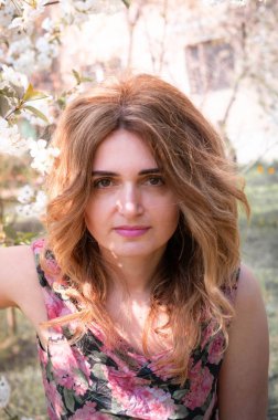 Güzel bir kadın, uzun saçlı, çiçek açan dalların yanında beyaz çiçekler ve yeşil yapraklarla bahçedeki güneş ışığıyla aydınlatılmış kameraya bakıyor.
