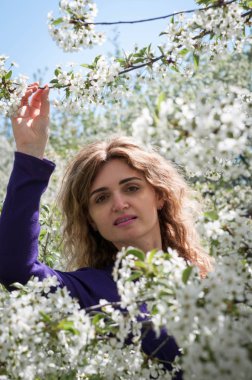 beautiful confident woman between white flowers on tree looking at camera and holding blossoming branch in garden in front of blue sky