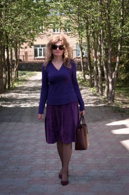 Mor etekli, bluzlu, uzun saçlı, yeşil ağaçların yanında çantalı güneş gözlüklü, parktaki kameraya bakan kendine güvenen güzel bir kadın.