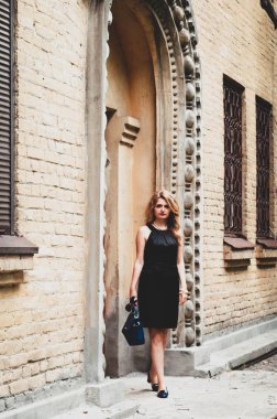 Çekici, kendine güvenen, uzun saçlı, zarif, küçük siyah elbiseli bir kadın. Elinde çanta, kaldırımda kemeri olan güzel sarı binanın yanındaki kameraya bakıyor.