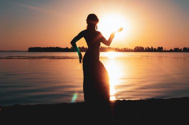 Sıska bir kadının karanlık, parıldayan silueti gün doğumunda nehrin yanındaki kırmızı gökyüzünün önünde gün ışığıyla aydınlanıyor.