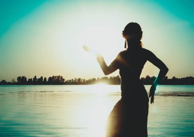 Sıska bir kadının karanlık, parıldayan silueti gün doğumunda nehrin yanındaki gökyüzünün önünde gün ışığıyla aydınlanıyor.