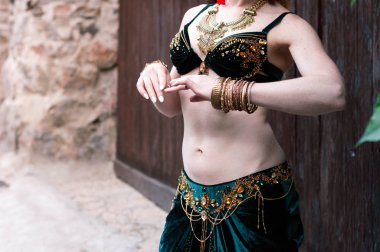 partial view of trible slim dencer woman in green folk costume standing near brick wall and brown wooden gates
