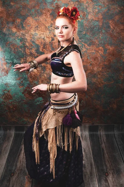 side view of beautiful woman in tradition tribal costume with vest, skirt and red floral circlet of flowers looking at camera in front of teal and orange background