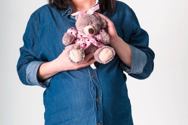 Mavi kot gömlekli hamile bir kadının elinde gri arka planda göğsüne yakın pembe elbiseli kahverengi oyuncak ayı oyuncağı vardı.