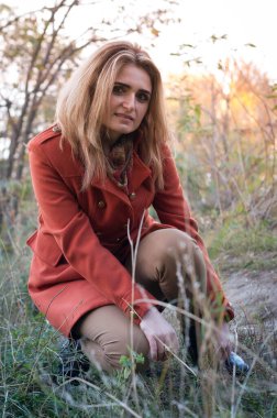 Sarı uzun saçlı, turuncu ceketli çekici bir kadın parkta kırmızı yapraklı çalıların önündeki kameraya bakıyor.