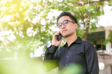 Mutlu genç yakışıklı adam stand altında açık havada ağaç ve iş için smartphone kullanarak konuşmak, gözlük adam