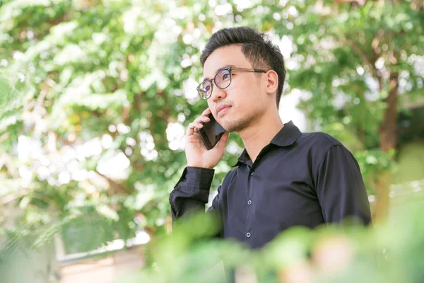 Happy young handsome man stand under tree outdoors and using smartphone for business talk, Glasses man