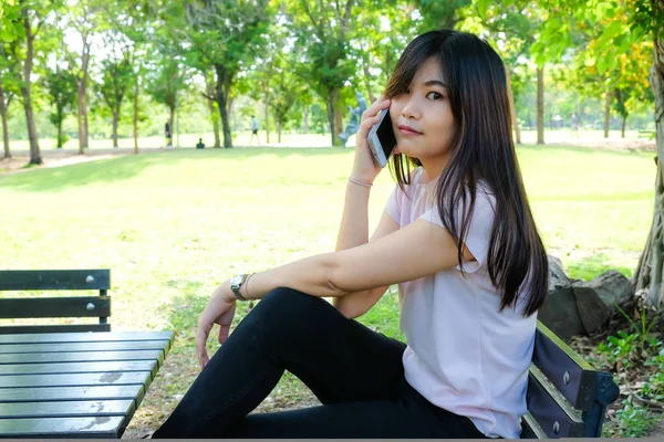 美丽的年轻亚洲妇女使用手机的社交媒体和互联网坐在公园长椅上 — 图库照片