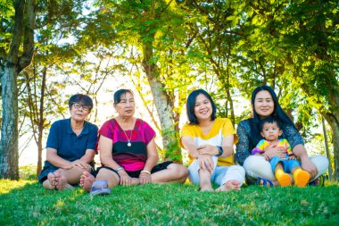 Üç kuşak Asyalı büyükanne, büyükbaba, torun yeşil çimenlerde oturuyorlar günbatımı aydınlığı açık hava parkı mutlu bir hayat.