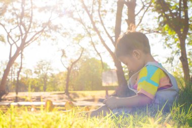 Ağaçların altında oturan sevimli Asyalı çocuk parkın gün batımında öğrendiği tablet bilgisayar teknolojisini kullanıyor.