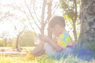 Şehir parkında ağacın altında oturan sevimli çocuk yeşil çimen gün batımında tablet bilgisayar kullanıyor.
