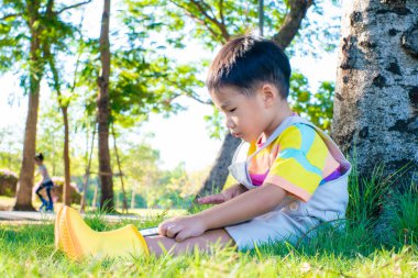 Şehir parkında ağacın altında oturan sevimli çocuk yeşil çimen gün batımında tablet bilgisayar kullanıyor.
