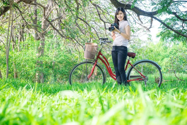 公园里骑红色自行车放松的妇女在绿地下使用相机 — 图库照片