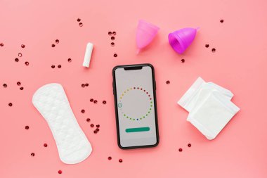 Poşetli regl kupası, hijyenik pedler, tampon, konfetili pembe arka planda adet takvimi olan cep telefonu. O dönemde alternatif kadın hijyen ürünü. Kadın sağlığı kavramı. 