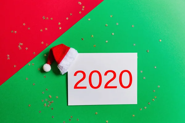 赤とコンフェッティで緑の背景にサンタの帽子と紙のブランク。クリスマスと新年のコンセプト。グリーティングカード、クリスマスお祝い2020 。フラットレイ、テンプレート、トップビュー、コピースペース、モックアップ — ストック写真