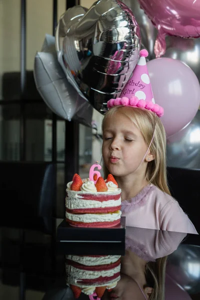 Indoor shot de niña bastante alegre con el pelo rubio soplando la vela, celebrar 6 años de edad cumpleaños, usar vestido de moda, tienen expresiones emocionadas. Concepto de infancia feliz —  Fotos de Stock