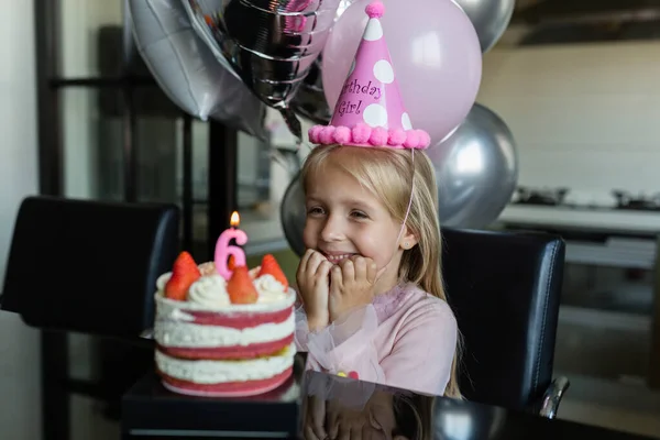 Indoor shot de niña bastante alegre con el pelo rubio soplando la vela, celebrar 6 años de edad cumpleaños, usar vestido de moda, tienen expresiones emocionadas. Concepto de infancia feliz —  Fotos de Stock
