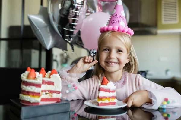 Indoor shot of pretty joyful little girl with blonde hair blowing out the candle, celebrate 6 years old birthday, wear fashionable dress, have excited expressions. Happy childhood concept — Stock Photo, Image