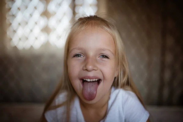 Jolie petite fille aux cheveux blonds montre la langue. Enfant avec un drôle de visage à la maison. Concept d'enfance heureuse — Photo