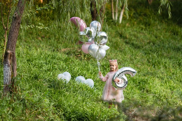Stylisches kleines Mädchen mit blonden Haaren, das im Park steht und Geburtstagsballons in der Hand hält. Sechs-Jahres-Feier. modisches Kind. Glückliche Kindheit — Stockfoto