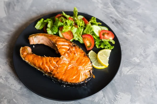 Fried salmon fish steak with vegetable salad and lemon on black plate. Creative layout made of fish, lettuce, tomato, cucumber, top view, flat lay, mockup, overhead. Healthy food concept