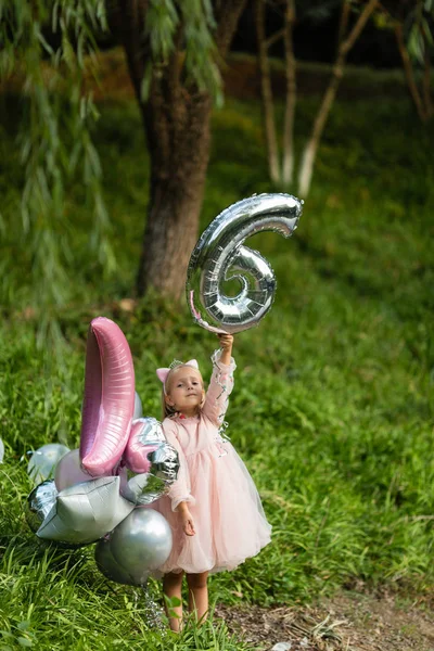 Foto al aire libre de niña bastante alegre con el pelo rubio celebrando 6 años de edad cumpleaños con globos, llevar vestido de moda, tienen expresiones emocionadas. Recuerdos de infancia feliz —  Fotos de Stock