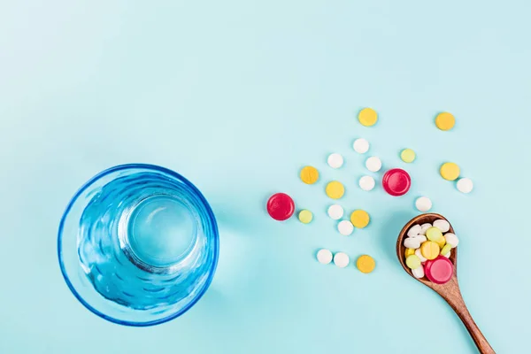 Pastillas de colores en cuchara de madera y un vaso de agua sobre un fondo azul pastel. Colocación plana, vista superior, techo, maqueta, plantilla. Farmacia y concepto médico — Foto de Stock