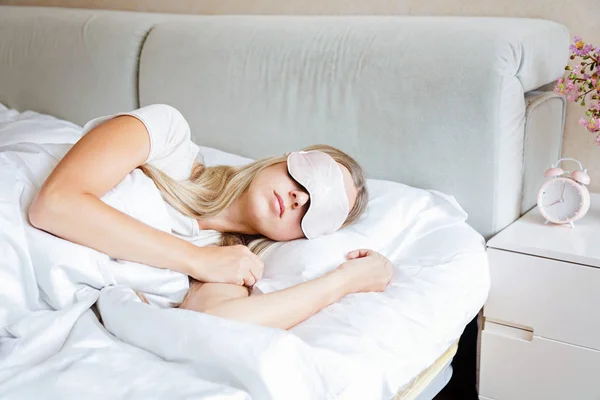 Young woman with long blonde hair sleeps on comfortable bed in a mask for sleeping. Blindfold on eye. Morning at home. White pillow and Blanket. Bedroom vibes