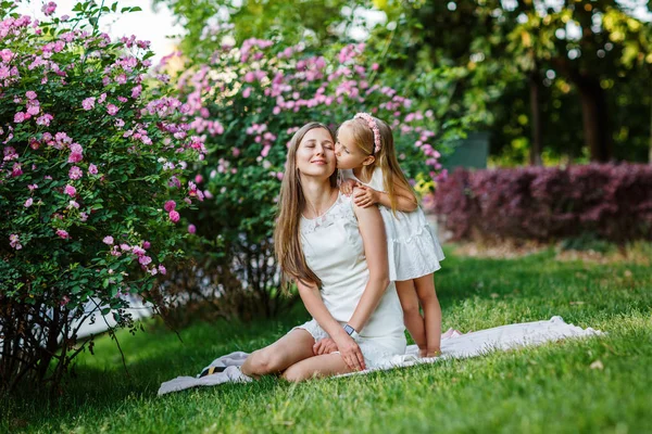 Portret van een mooie vrouw en meisje buiten. Klein meisje dat haar lieve moeder kust in het zomerpark met bloeiende bloemen. Gelukkige Moederdag concept — Stockfoto
