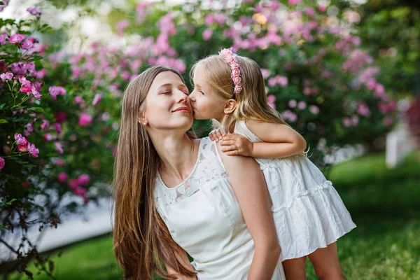 Retrato de mujer y niña hermosa al aire libre. Niña besando a su encantadora madre en el parque de verano con flores en flor. Concepto del día de la madre feliz — Foto de Stock