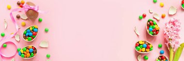 Çikolatalı Paskalya yumurtası ve pastel pembe kağıtta renkli şekerler. Düz konum, üst görünüm, üstten görünüm, kopyalama alanı, şablon, model. Web tasarımı için yatay pankart.