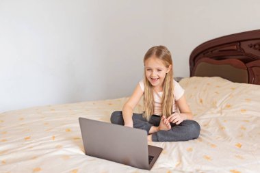 Evde kalıp koronavirüs salgınını önleyin. Yatakta oturan ve dizüstü bilgisayar kullanan sarı saçlı küçük kız. Önleme salgını. Teknoloji kullanan çocuk