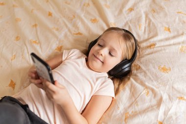 Evde kalıp koronavirüs salgınını önleyin. Kablosuz kulaklıkla yatakta yatan sarı saçlı küçük kız. Önleme salgını. Çocuk teknolojiyi kullanıyor. çevrimiçi eğitim.