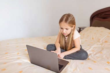 Evde kalıp koronavirüs salgınını önleyin. Yatakta oturan ve dizüstü bilgisayar kullanan sarı saçlı küçük kız. Önleme salgını. Teknoloji kullanan çocuk