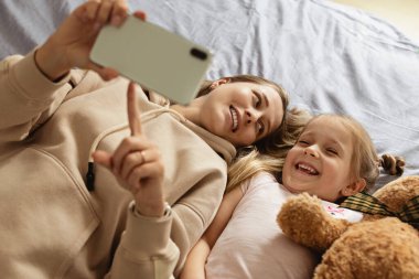 Anne ve kızı akıllı telefondan tek başına yatakta gülümseyen yüzüyle video izliyorlar. Evde mutlu yüzlü cep telefonu kullanan bir çocuk. Evde karantina Coronavirus COVID-19 pandemik önleme