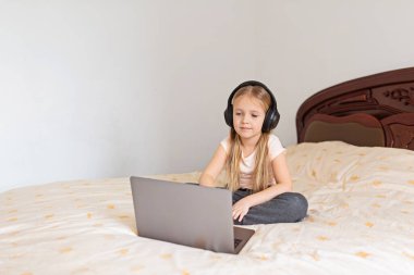 Evde kalıp koronavirüs salgınını önleyin. Yatakta oturan ve dizüstü bilgisayar kullanan sarı saçlı küçük kız. Önleme salgını. Teknoloji kullanan çocuk