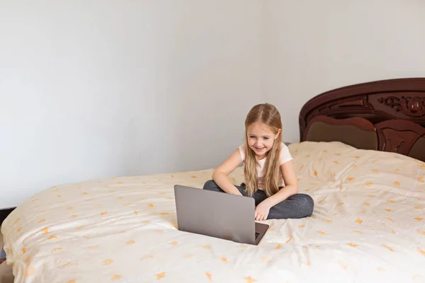 Stay at home quarantine coronavirus pandemic prevention. Little girl with blonde hair sitting on the bed and using laptop. Prevention epidemic. Child using technology